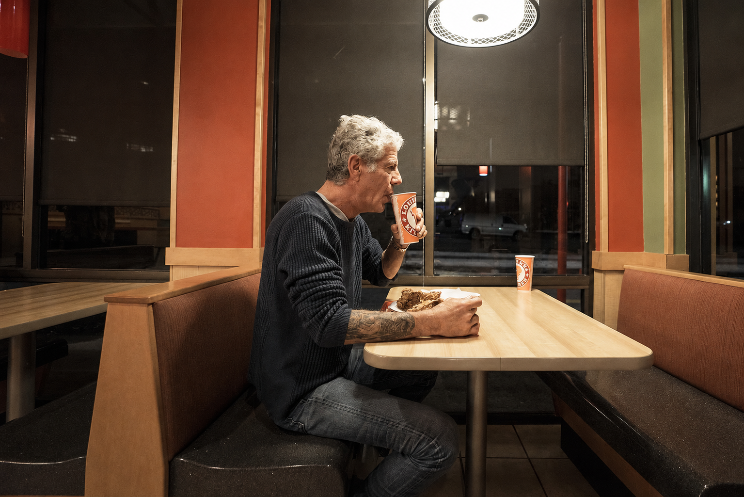 Anthony Bourdain seated inside a Popeyes restaurant with fried chicken and a drink